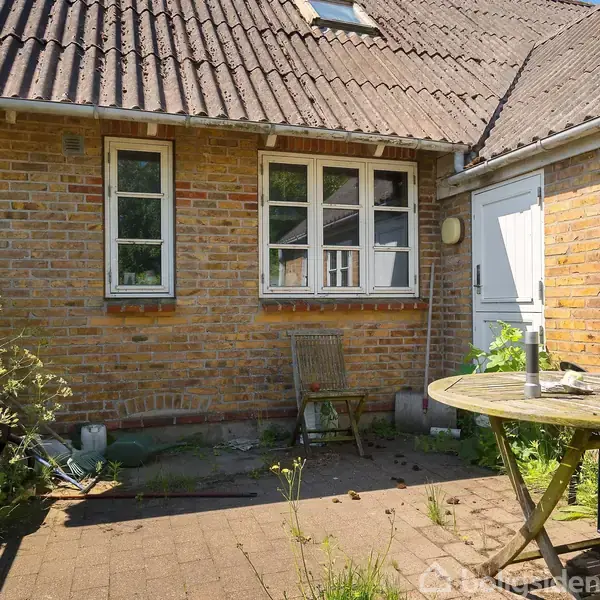 En rund træstol er placeret på en fliseterrasse foran et murstenshus med to vinduer. En stol står op ad væggen i haven.