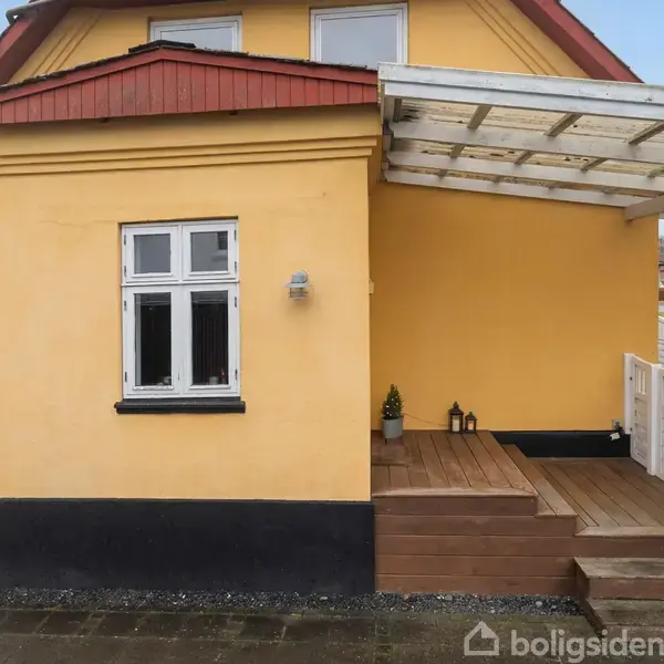 Et hus med gul facade står stille. Ved siden af er en overdækket træterrasse med trappetrin. En hvid havelåge og stakit omkranser området.