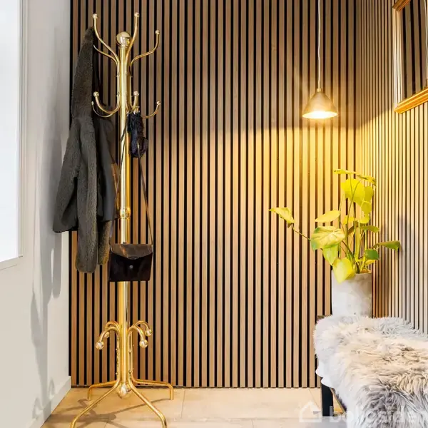 En gylden knagerække holder jakker i en entré med lodrette træpaneler. En plante i potte står ved en bænk med pelsdække under en væglampe, der kaster lys på væggen.