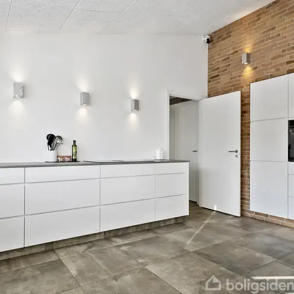 En garagebygning med to hvide porte står i midten, omgivet af en velplejet græsplæne og en belagt indkørsel. Baggrunden har klare himmel og hække.