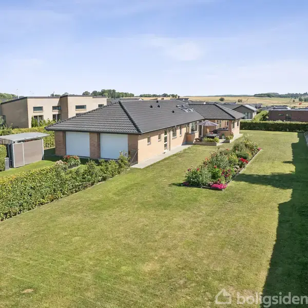 En lysebrun villa står omgivet af en blomstrende have med farverige blomster. Husets tag er mørkt, og himlen er klar og blå. Græsplænen er velplejet.