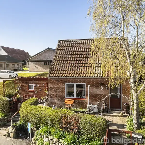 En charmerende murstenshus står på en velplejet have med en hæk og stensætning. Husnummer 8 er synligt. I baggrunden ses flere lignende huse og en parkeret bil.