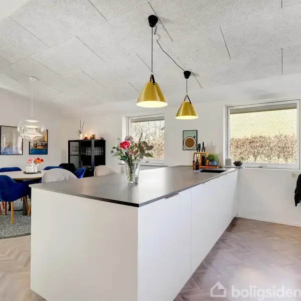 Køkken med hvidt moderne køkkenbord, blomster i vase, og to lysekroner over. I baggrunden ses spiseområde med blå stole og rundt tæppe. Vinduer viser buskadser udenfor.