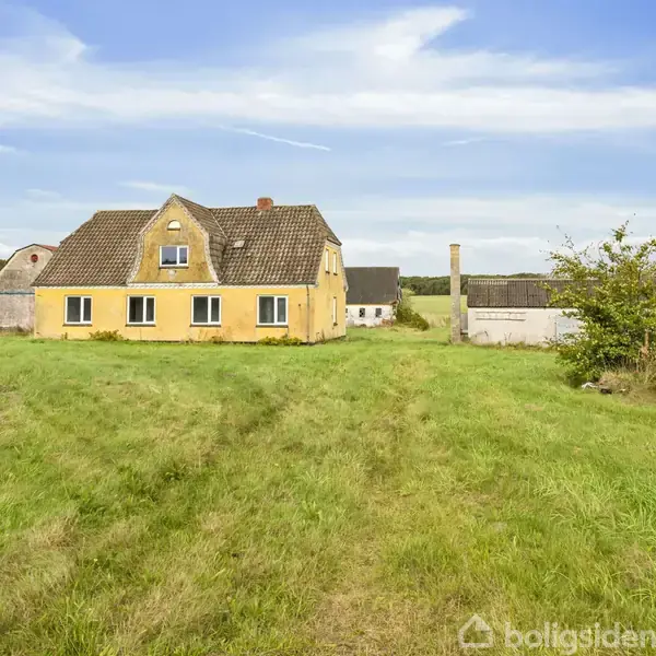 Gul murstensbygning med flere vinduer står stille på en stor græsplæne, omgivet af åbne marker og træer under en blå himmel.