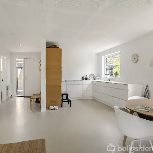 Køkkenet er lyst og rummeligt med hvide skabe og en bordplade langs væggen. Et rundt spisebord med stole står ved siden af et vindue. Ingen tekst er synlig.