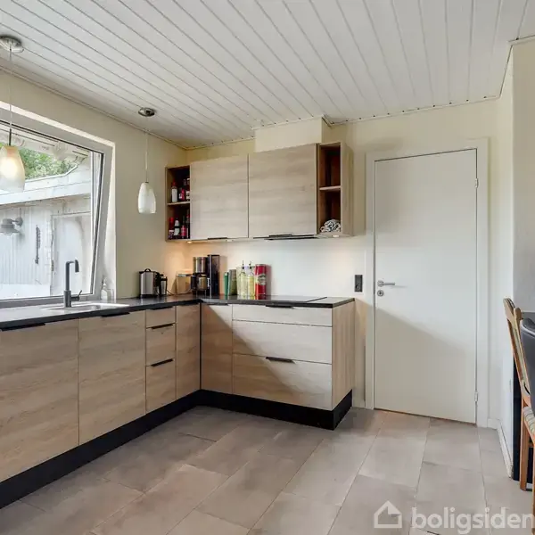 Moderne køkken med lyse træskabe og sorte bordplader. Køkkenet har integreret ovn og store vinduer til venstre, der giver udsigt til en terrasse. En dør til højre fører til et andet rum.