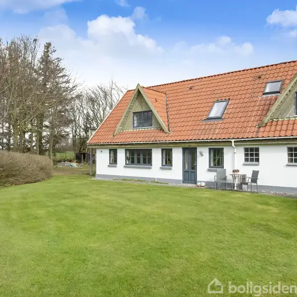 Et hvidt hus med rødt tegltag står i en velplejet have. Der er et par stole og et bord tæt på hoveddøren. Omkring er der træer og en blå himmel.