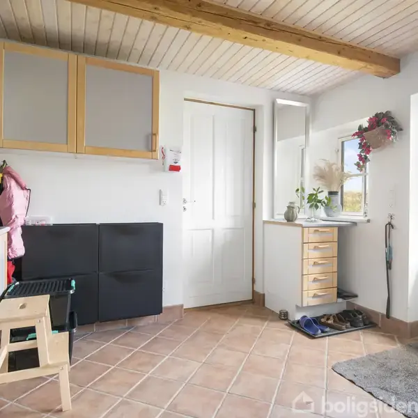 Et lyst bryggers med flisegulv og træloft. Skabe og hylder er monteret på væggene. En dør fører ud til haven. Vintertøj hænger på knager.