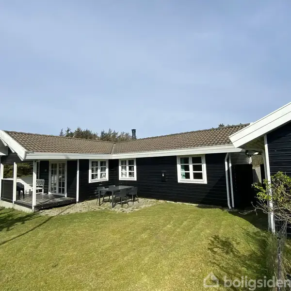 Et sort, træhus med hvide vinduer står i en grøn have. Et bord med stole ses foran, mens omgivende træer giver en naturlig baggrund.