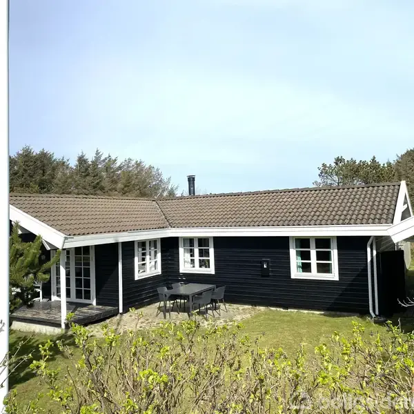 Sort træhus med hvide vinduer og brunligt tag. Huset står på en græsplæne omkranset af buske og træer. Foran huset står et bord med stole på en terrasse.