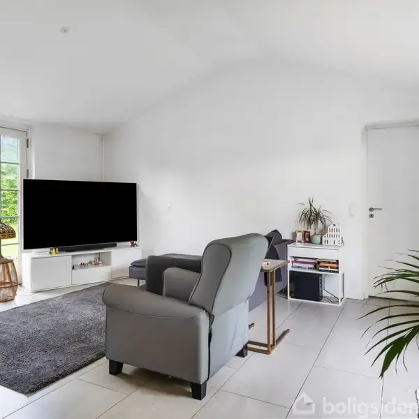 Et gråt sofaarrangement står i en lys stue med store vinduer til venstre. En fladskærms-tv ses foran. Grønne planter og en dør til højre skaber en hyggelig atmosfære.