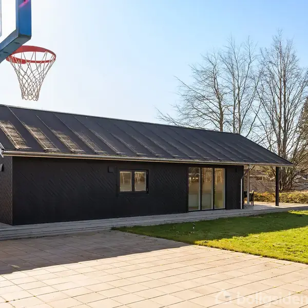 En sort bygning står stille på en flisebelagt terrasse ved siden af en grøn græsplæne. En basketballkurv er til venstre, omgivet af træer og klar himmel.