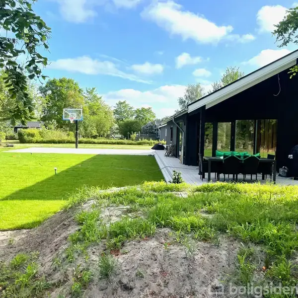 Et moderne hus i sort træ står i en frodig have med grønne plæner. En basketballkurv står på en flisebelagt terrasse, omgivet af træer og blå himmel.