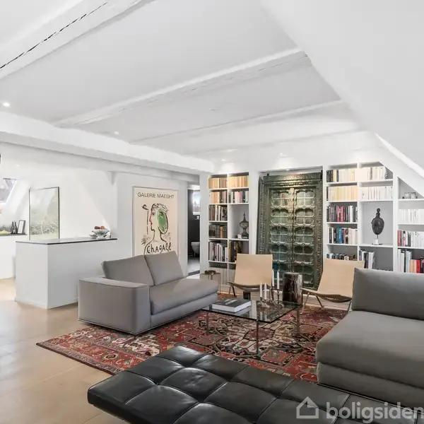 Stue indrettet med grå sofa og lænestole omkring et glasbord. Kunst og bøger pryder væggene. Stuemiljøet er lyst med store hylder og et kunstværk med teksten "GALERIE MOURLOT GRAPHIQUE."