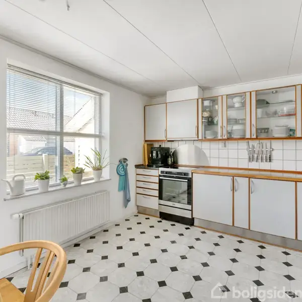 Et lyst køkken med hvide skabe og sort-hvide gulvfliser. Komfuret står ved væggen under glasskabene. En stor vindueskarm med planter giver udsigt til nabolaget udenfor.