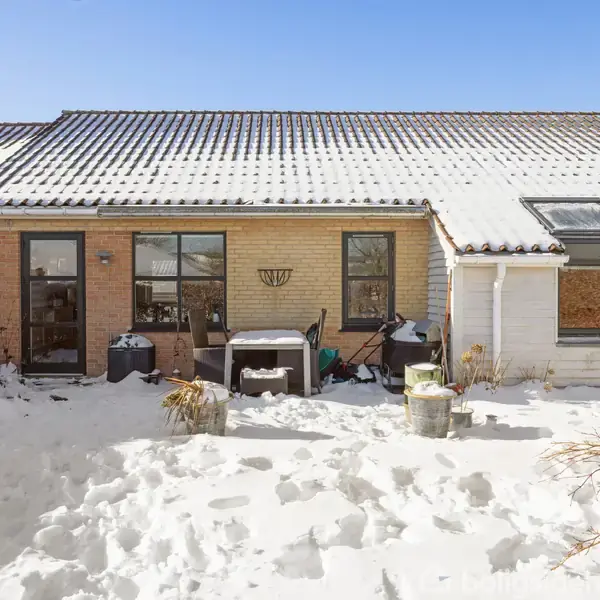 Hus med sneklædt have; terrassedør og vinduer i brun murstensbygning til venstre, hvid træbeklædning med vindue til højre. Sollys skinner på tag og omgivelser. Ingen tekst.