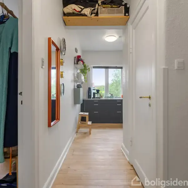 Gang med træplankegulv og hvidmalede vægge. En dør til højre, farvede kjoler hænger til venstre. For enden ses et køkken med grå skabe og et vindue.