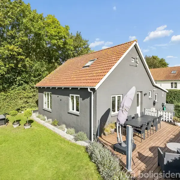 Et gråmalet hus med rødt tegltag står i en have. Huset har flere vinduer, og en terrasse med havemøbler og en parasol ved siden. Græsplæne og træer omkranser huset.