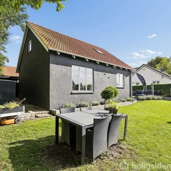 Sort hus med rødt tag står i en frodig have. Foran huset er et spisebord med stole og en fuglefodringsstander. Området er omkranset af hække og træer.