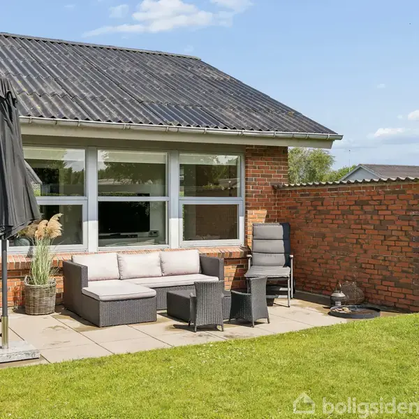 Terrassemøbler står på en flisebelagt terrasse ved et rødt murstenshus. En parasol er slået op ved siden af sofaen, og der anes en velplejet græsplæne under en blå skyfri himmel.