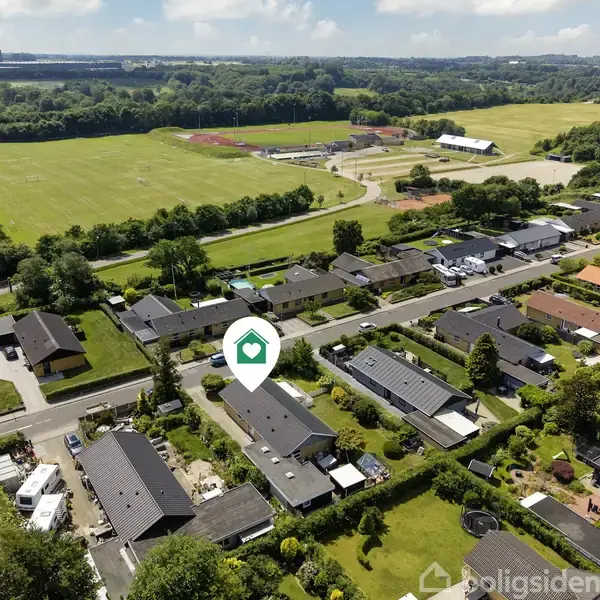 Et parcelhuskvarter med en grøn mark i baggrunden. En hus-ikon-markeret ejendom ligger blandt andre huse med velplejede haver, belyst af dagslys under en blå himmel med få skyer.