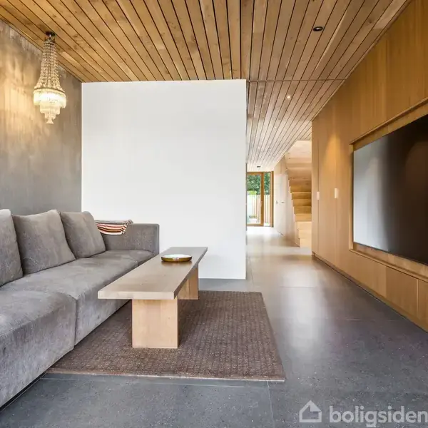 En lang grå sofa står mod en betonvæg med en moderne trælampe, mens en fladskærm er indbygget i trævæggen. Rummet har et træloft og åben passage.