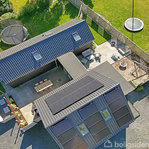 Hus med solpaneler på taget er omgivet af en have med en trampolin. Der er en terrasse med bord og stole samt en grusindkørsel.