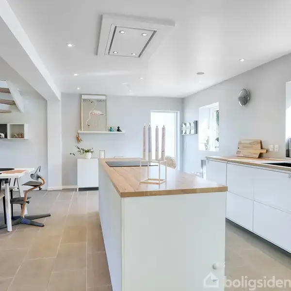 Køkkenet har en stor, hvid køkkenø med træoverflade. Omkring er spisebord med stole nær en trappeopgang. Lyse vægge giver et moderne, lyst udseende med minimalistisk indretning.