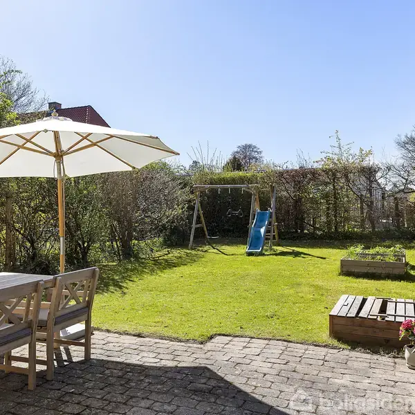 Terrassebord med parasol står på en stenbelagt terrasse. I baggrunden er der en græsplæne med en gynge og rutsjebane samt blomsterkasser. Sollys dækker haven.