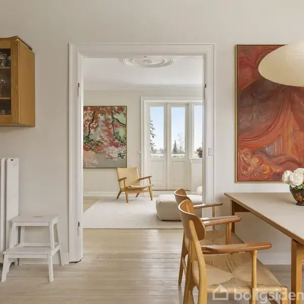 Et moderne, lyst køkken med træbord og stole leder ind til en stue med to malerier på væggene. Stuen har et stort vindue, der lader lys strømme ind.
