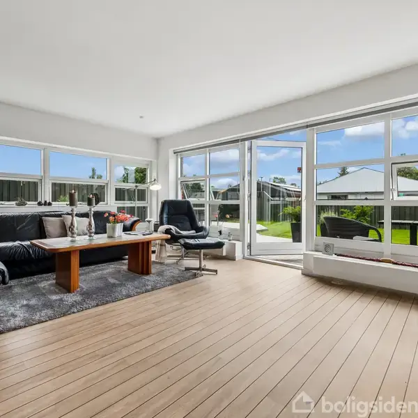 Stue med sort sofa og bord på gråt tæppe, omgivet af store vinduer med udsigt til en grøn have. Et sort lænestolsæt står ved siden af, i en lys og rummelig atmosfære.