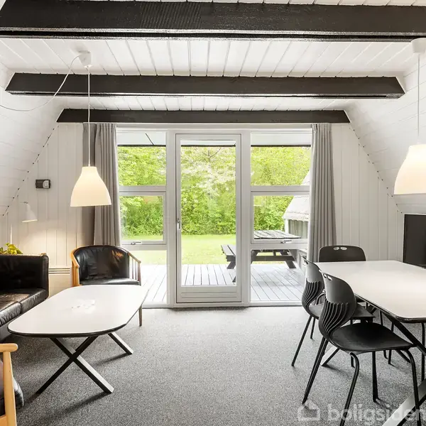Stue med sort lædersofa og spisebord med stole, belyst af pendellamper. Store glasdøre åbner til terrasse med udsigt over grøn have. Hvidmalede vægge og synlige bjælker.