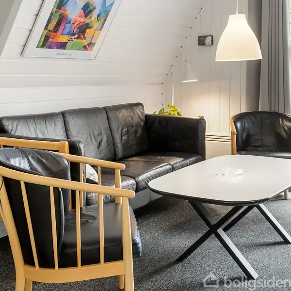 Sort lædersofa og to stole står omkring et rektangulært sofabord i en lys stue med skråloft. Lampe hænger over bordet. Maleri og stort vindue til højre.
