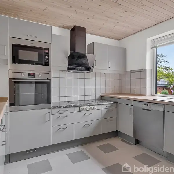 Et moderne køkken med hvide skabe, indbygget ovn, mikroovn og kogeplade. Rummet er lyst med en træloft og stort vindue med udsigt til en have.