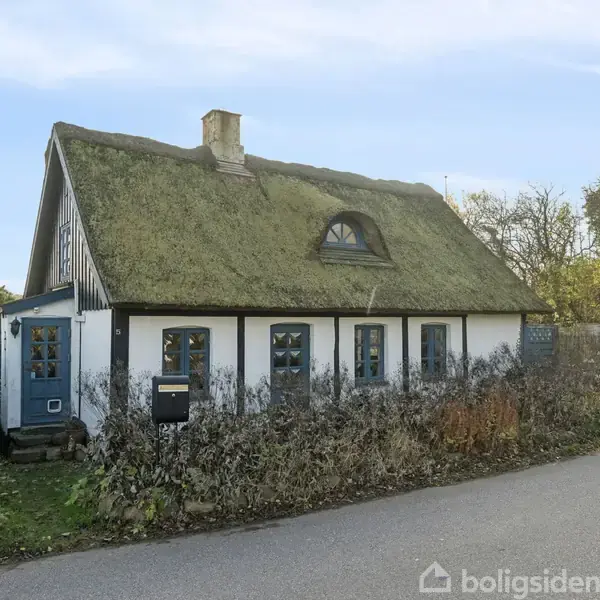 Stråtækt hus står stille på en landsvej, omgivet af frodig vegetation. Det har blå dør, småsprossede vinduer og en skorsten, hvilket giver et idyllisk og landligt udtryk.