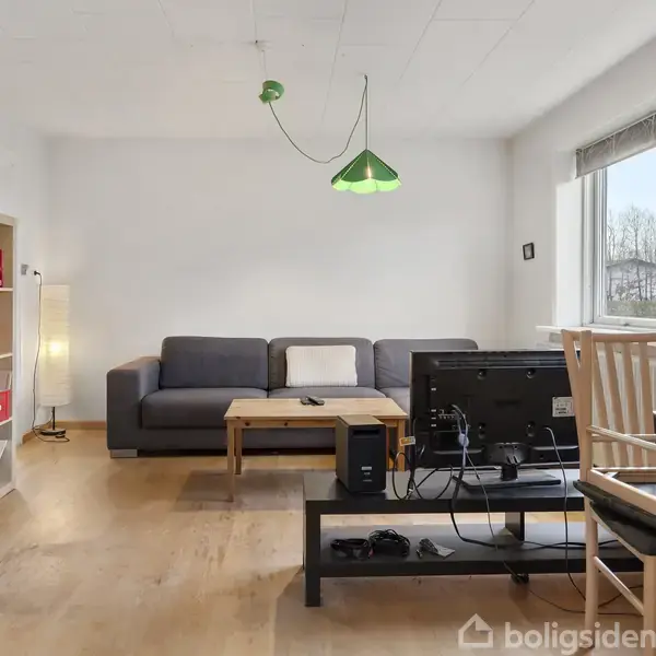 Sofa står i stuen med et sofabord foran. Et grønt lys hænger over, og en reol fyldt med bøger står til venstre. Et vindue er til højre. En stol står oprejst på et bord med en skærm.