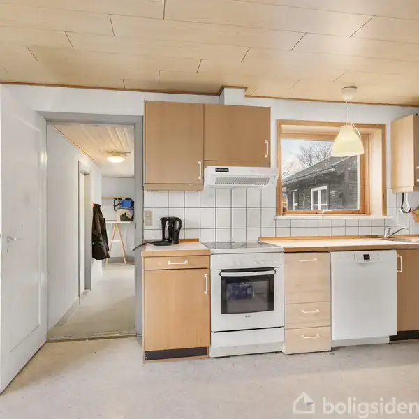 En køkkenindretning med træskabe og hvide fliser, omfatter en komfur og en opvaskemaskine. Rummet er lyst med store vinduer, og en åben dør leder til en gang.