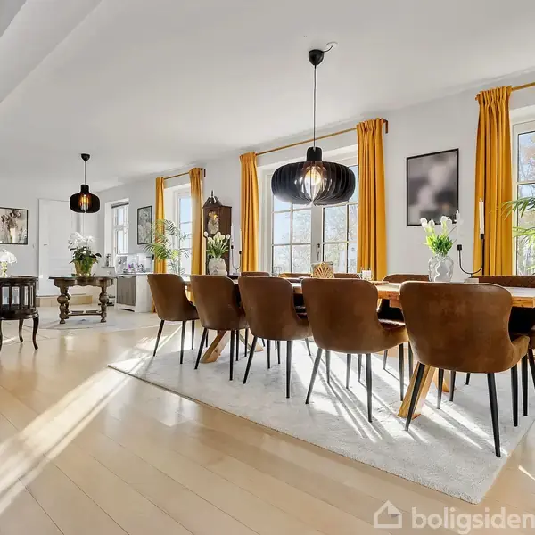 En lang spisebord med brune stole står i et solbeskinnet rum med store vinduer og tykke, gule gardiner. Der er planter og lysekroner, hvilket skaber en elegant indretning.