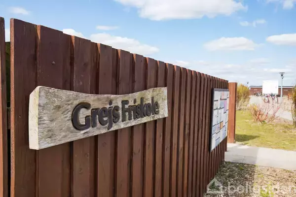 Træhegn med skiltet "Grejs Friskole" langs en vej i et åbent område med blå himmel og skyer i baggrunden.