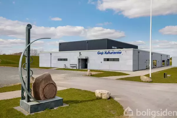 En grå bygning, Grejs Kulturcenter, står omgivet af grønne områder og en skulptur, der består af sten og metalbuer, placeret i forgrunden under en blå himmel med spredte skyer.