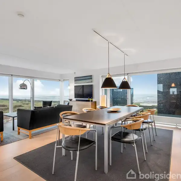 Et moderne spisestuemiljø med et langt bord og otte stole. Tre lamper hænger over bordet. Store vinduer giver havudsigt, og en stue med sofaer ses i baggrunden.