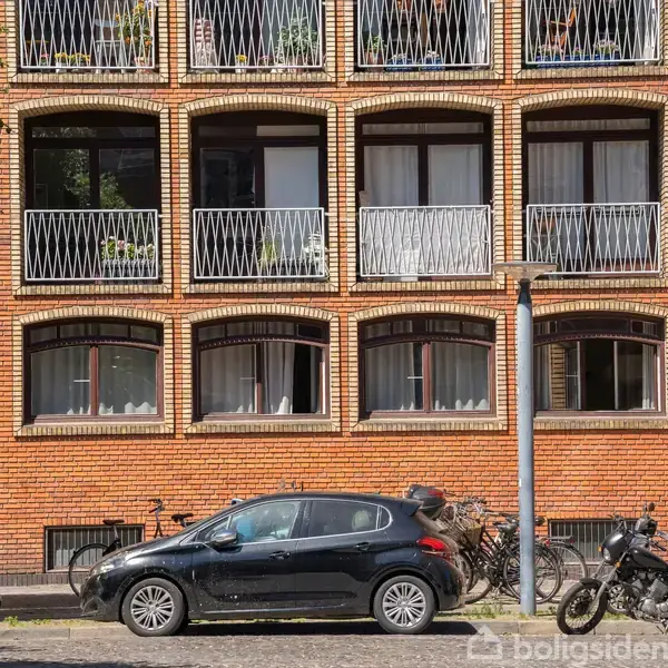 En sort bil holder parkeret foran en rødstensbygning med buede vinduer og altaner. Cykler er parkeret ved siden af bilen. Træer kaster skygger på gaden.