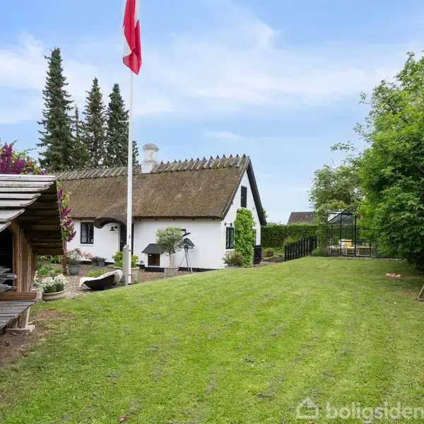 Et hvidt, stråtækt hus står i en have med en flagstang, flag, og græsplæne. Til venstre er der en træoverdækning, og til højre en gynge.
