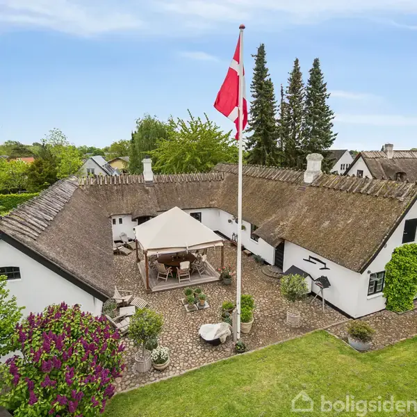 Bindingsværkshus med stråtag omgivet af grøn have. I gårdhaven står en pavillon og et flagstang med Dannebrog. Omgivet af andre huse og grønne træer under en blå himmel.