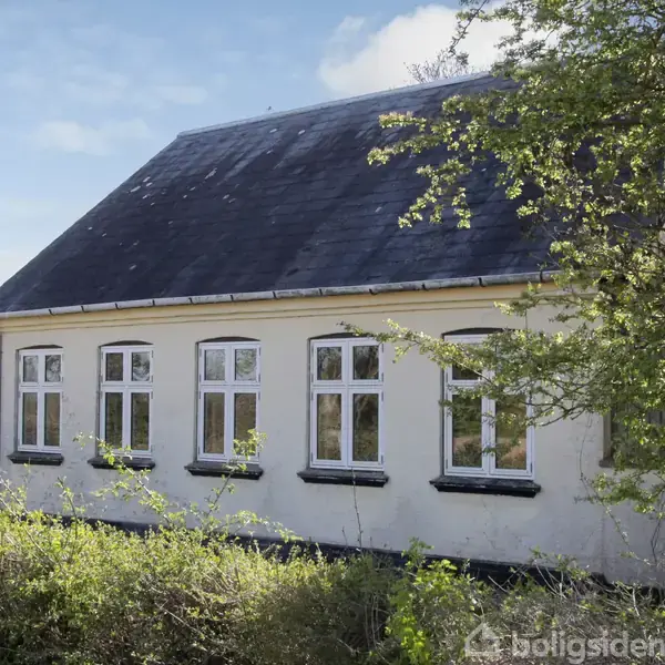 Gult hus stående stille i en frodig have med græs og buske omkring, lyseblå himmel bagved.