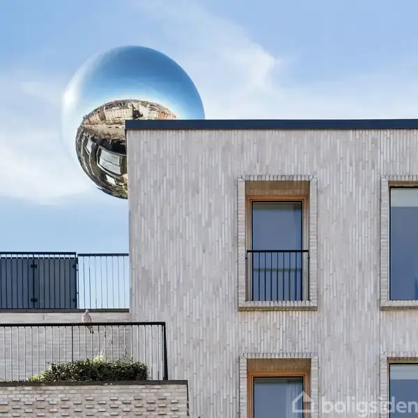 Bygning med hvide mursten og store vinduer, der har altaner med sorte gelændere. En stor, reflekterende kugle svæver i baggrunden på en klar blå himmel.