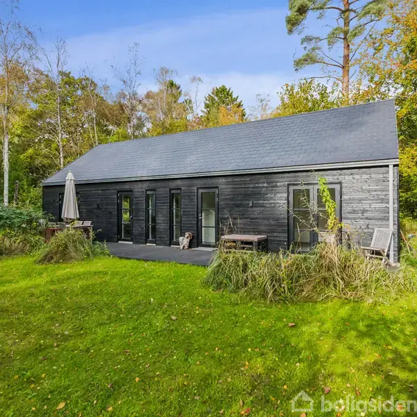 Et sort træhus står på en grøn græsplæne. Huset har store vinduer og en terrasse med parasol. Omgivet af høje træer og buske, fremstår området fredeligt og naturligt.