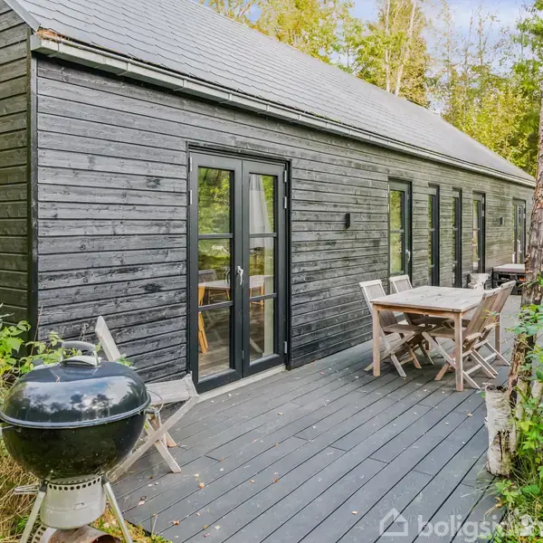 Et sort træhus står i en skovklædt have. En udendørs terrasse har havemøbler og en grill. Naturen omkring skaber en rolig atmosfære med træer og buske.