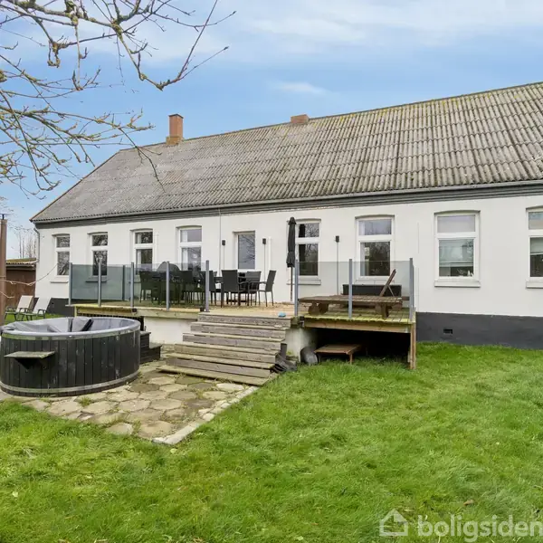 Et hvidt enfamilieshus med gråt saddeltag står i en grøn have. Trædæk med havemøbler foran huset. En rund, sort udendørs spa ved siden af trappen.