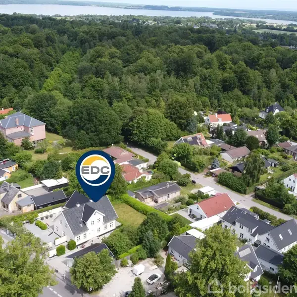 Et hus markeret med et EDC-skilt ses fra oven. Det står i et grønt, skovklædt boligområde med flere lignende bebyggelser rundt om.